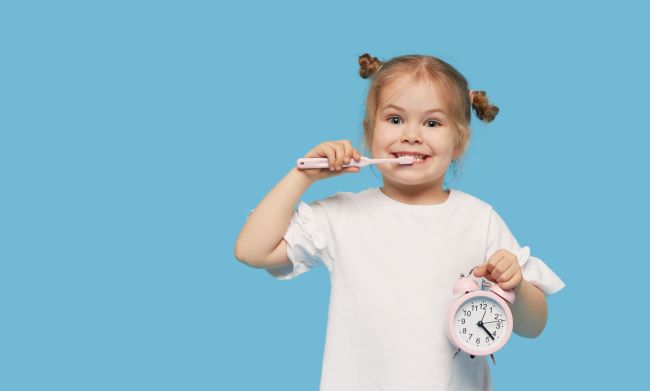 Malá blonďatá holčička v bílém tričku drží v jedné ruce růžové budíkové hodiny a ve druhé má zubní kartáček, myje si zuby a usmívá se.